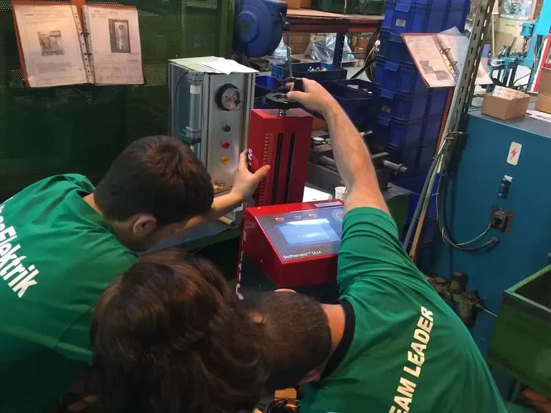 Fabrika ortamında kırmızı Dotpeenator SA14 markalama cihazını ayarlayan yeşil tişörtlü çalışanlar.