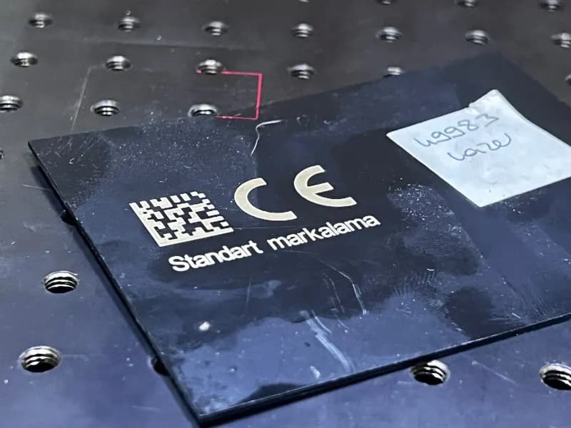 Mat siyah plastik parça üzerine 20W fiber lazer ile DataMatrix kodu, CE sembolü ve metin içeriği markalama operasyonu.